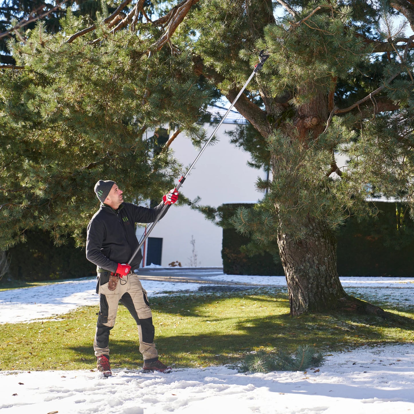 Man using FELCO Pole Pruner 290-4M to prune tree branches in a snowy landscape.